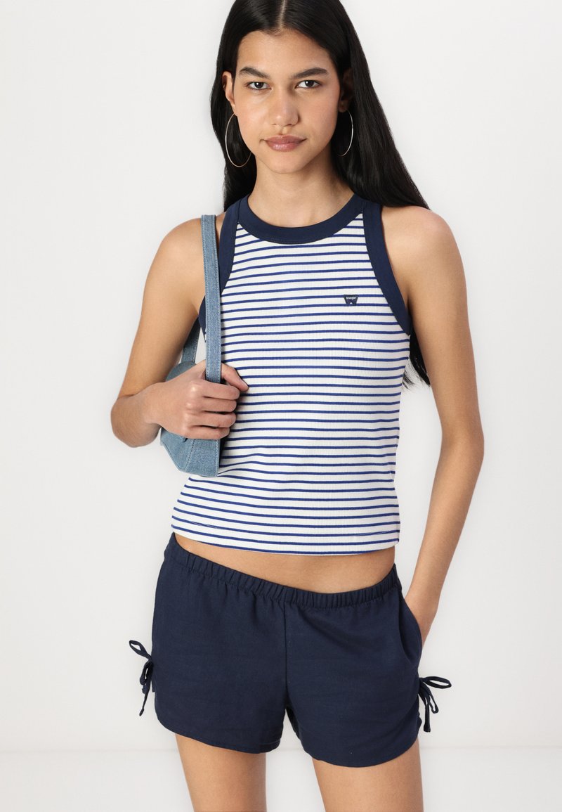 Top de tirantes a rayas en blanco y azul marino con un escote redondo, acompañado de pantalones cortos azules marinos con lazos laterales. El modelo sostiene un bolso de mezclilla.