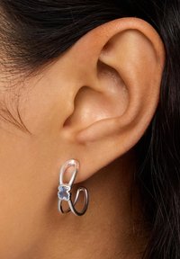 Srebrna kolczyk kołowy z zakrzywionym wzorem, ozdobiony niebieskim kamieniem szlachetnym na środku i gładką, wypolerowaną powierzchnią.