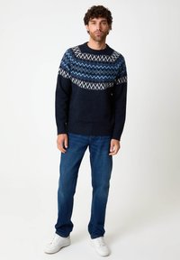 Dunkelblauer Strickpullover mit geometrischen Mustern in Hellblau und Weiß auf der Brust, kombiniert mit dunkelblauen Jeans und weißen Sneakers.