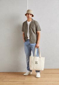 Camisa gris de manga corta, camiseta blanca, jeans de color azul claro, sombrero tipo cubo beige y una bolsa de tote color crema con texto negro, de pie sobre un suelo de madera.