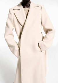 Beige wool coat with a structured design, wide lapels, side pockets, and a soft texture, featuring a wrap closure across the front.