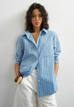 Kvinde med krøllet mørkt hår iført en løs blåstribet skjorte med knapper og hvide bukser, stående mod en enkel lys væg.