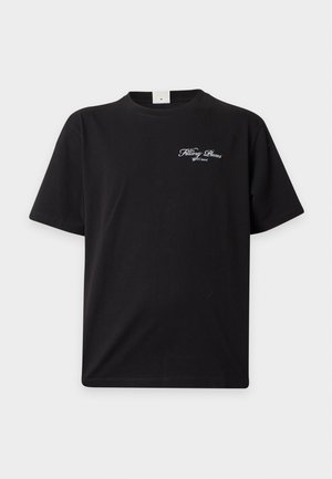 Jeune homme debout, face à l'avant, portant un T-shirt noir à manches courtes, un jean bleu droit et des baskets noires sur un fond blanc uni.