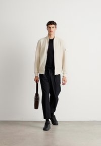 Giovane uomo che cammina in avanti indossando una giacca crema, una camicia nera, pantaloni neri e scarpe nere, con una valigetta marrone nella mano destra.