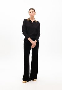 Schwarzer Pullover mit strukturiertem, gold-aczentuiertem Kragen, kombiniert mit weiten schwarzen Samthosen und goldenen Schuhen, vor einem weißen Hintergrund platziert.