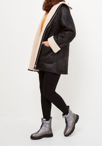 Abrigo de piel de oveja negro con forro beige y grandes bolsillos, combinado con botas metálicas plateadas hasta el tobillo y leggings negros.