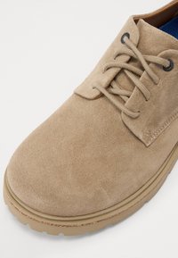 Beige semsket lissesko med rund tå, flat såle og svarte øyer. Har en sydd søm langs sålen for ekstra detaljer.