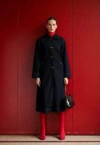Manteau en denim avec un col, cinq boutons, poches latérales, associé à des collants rouges et des talons. Portant un sac noir avec un motif texturé.
