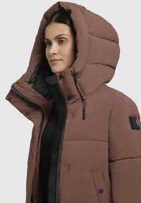 Barna puffasztott kabát nagy kapucnival, fekete béléssel, cipzárral és patenttal záródik, valamint bal vállon jelvény részlet található. Simá szövet.