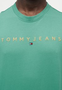 Πράσινο βαμβακερό T-shirt με στρογγυλή λαιμόκοψη, διακοσμημένο με κίτρινη κεντημένη επιγραφή "TOMMYJEANS" και μικρό κόκκινο, λευκό και μπλε λογότυπο κάτω από το κείμενο.