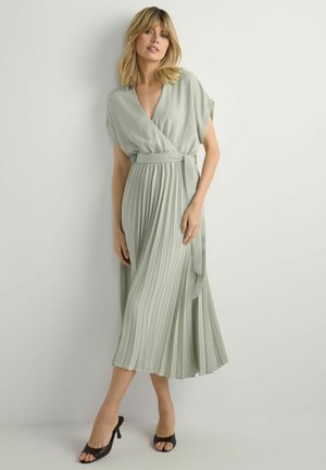 Kobieta o blond włosach do ramion w jasnozielonej plisowanej sukience kopertowej i czarnych butach na obcasie z odkrytymi palcami, stojąca na tle jasnej ściany.