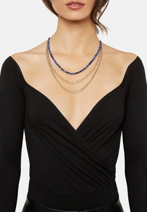 Gelaagde ketting met drie strengen: donkerblauwe kralen, gouden schakelketting, en een delicate gouden ketting; gedragen over een zwarte wikkeltop.