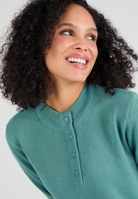 Türkis gestrickter Pullover mit rundem Kragen, der eine Knopfleiste und strukturierte Rippdetails am Hals und den Bündchen aufweist. Weicher, gemütlicher Stoff.