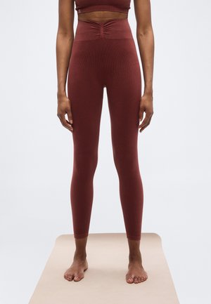 Persona de pie descalza sobre una esterilla beige, vistiendo leggings de talle alto color granate y un top corto a juego, con fondo liso.