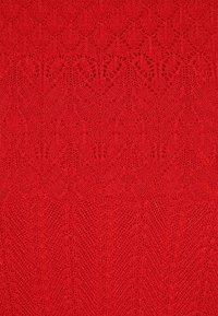 Rote gestrickte Stoff mit filigranen Mustern, offenen Ösen und strukturierten Details, der eine Mischung aus geometrischen und floralen Formen zeigt.