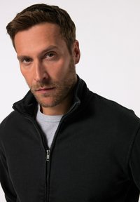 Chaqueta negra con cremallera y cuello alto, con una cremallera plateada, usada sobre una camiseta gris. Textura de tela suave y diseño casual.
