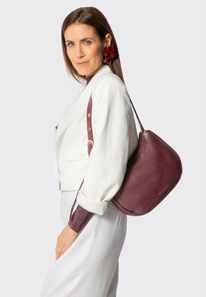 Vrouw draagt een wit gestreept colbert en broek, met een grote donkerrode leren schoudertas en bijpassende oorbel in bloemdessin.