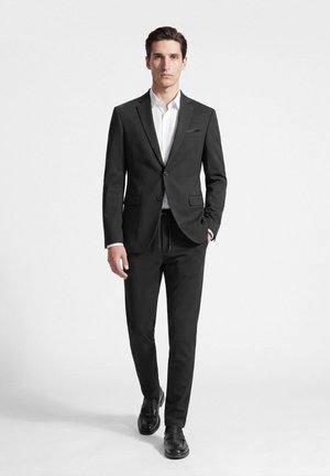 Mann geht vorwärts und trägt eine schwarze Zweiknopf-Anzugsjacke mit passenden Hosen und schwarzen Schuhen, darunter ein weißes Hemd, eine Hand in der Tasche.