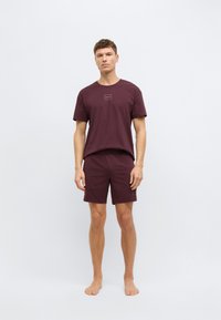 Σετ από βαμβακερό t-shirt και σορτς σε μαρονοκόκκινο χρώμα, με λογότυπο στο στήθος, κοντά μανίκια και casual εφαρμογή, συνοδευόμενο από γυμνά πόδια.