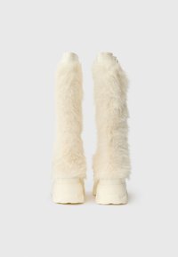 Knee-high boty vyrobené z nadýchaného krémového syntetického kožešiny, s masivní bílou podrážkou. Mají hladký povrch nahoře a texturované tělo.