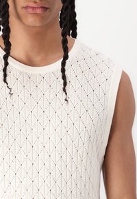 Strikket ærmeløs top i off-white med et struktureret diamantmønster, rund halsudskæring og åbent væv for åndbarhed.