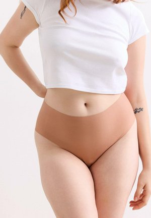 Frau trägt ein weißes, kurz geschnittenes T-Shirt und nahtlose beige High-Waist-Unterwäsche, wobei der Bauchbereich und Tattoos an beiden Armen sichtbar sind.
