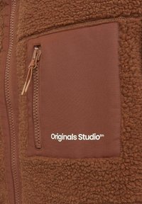 Brązowa dzianina polaru z gładką łatką tekstylną, wyposażona w kieszeń na zamek błyskawiczny oraz logo "Originals Studio™" w białym kolorze.