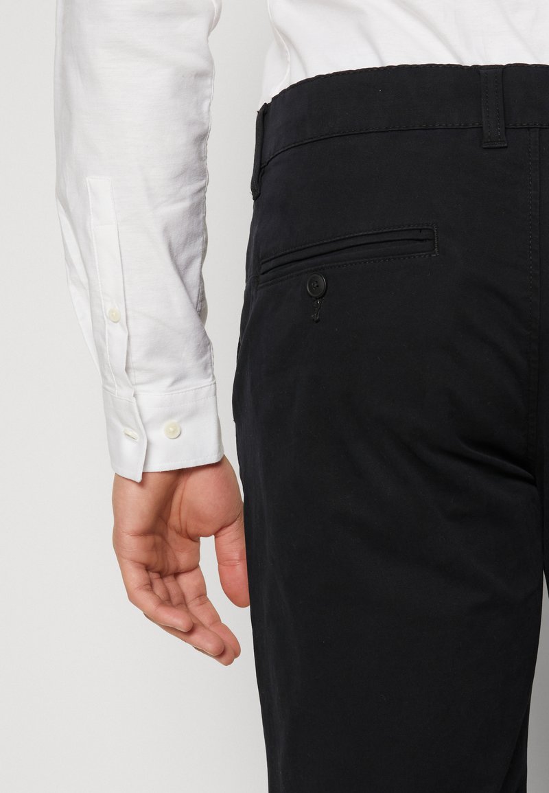 Main d'homme détendue sur le côté, portant une chemise blanche et un pantalon noir avec la poche arrière boutonnée.