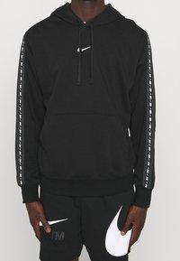 Persona que lleva puesta una sudadera negra de Nike y pantalones cortos a juego, con logotipos en blanco de swoosh de Nike y franjas de marca en las mangas.