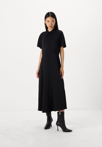 Schwarzes, kurzärmliges Kleid mit Kragen, fließender A-Linie, glatter Textur und verdeckten Seitentaschen. Kombiniert mit schwarzen Stiefeletten.