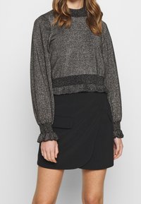 Kvinna klädd i en metallic grå långärmad kort tröja med volangärmar och en svart inskriven minikjol med fickor.