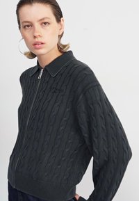 Donkergroene ritstrui met kabeltricot textuur, geribbelde manchetten en kraag. Bevat het merkje op de borst. Casual pasvorm.