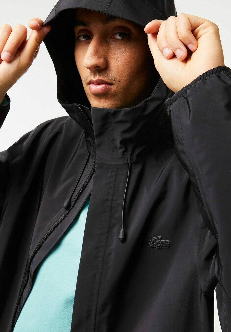 Homme ajustant la capuche d'une veste zippée noire avec un petit logo sur la poitrine, portant une chemise bleu clair en dessous, sur un fond uni.