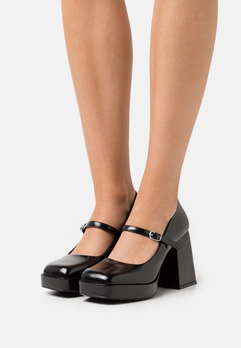 Jambes portant des escarpins Mary Jane à plateforme en cuir verni noir, avec des talons blocs épais et des brides à la cheville avec boucles argentées.