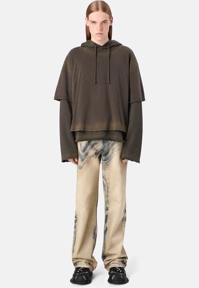 Felpa oversize verde oliva con cappuccio e cordoncino, indossata sopra una maglietta a maniche lunghe. Abbinata a jeans larghi a righe color beige chiaro e sneakers nere.