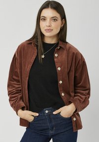 Veste-chemise en velours côtelé marron avec une fermeture à boutons, dotée d'une coupe décontractée et de grandes poches, portée sur un pull noir.