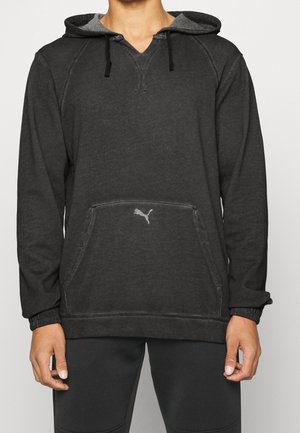 Homme portant un sweat à capuche Puma gris foncé avec poche avant et cordons noirs, associé à un pantalon noir, sur fond uni.