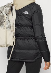 Personne portant une doudoune noire avec le logo The North Face, une écharpe beige, et un sac à dos beige à motif camouflage porté sur une épaule.
