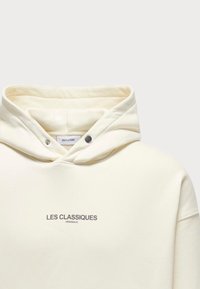 Hoodie crème avec cordon de serrage, doté d'une poche frontale et d'un texte imprimé "LES CLASSIQUES" en gris. Tissu doux avec un design décontracté.
