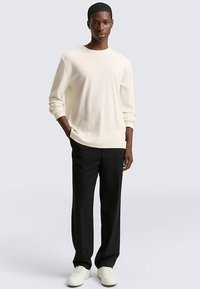 Light cream long-sleeve shirt, crew neckline, relaxed fit, paired with black pants and white sneakers, set against a neutral background.