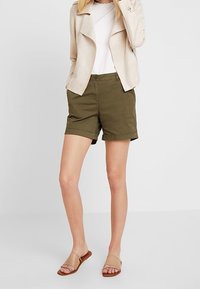 Shorts verts kaki à coupe décontractée, associés à un haut blanc et une veste beige clair texturée. Des sandales à semelle plate complètent le look.