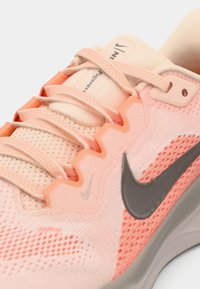Gros plan sur une chaussure de course Nike rose avec des accents corail, un logo swoosh gris et un tissu en maille texturée avec des lacets noués sur la languette.