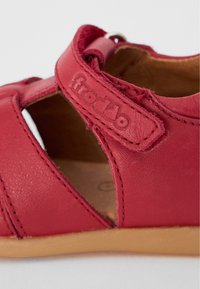 Rød læder sandal med lukket bagside, åbne sider og syning detaljer. Har en rem med et logo og en tan gummisål.
