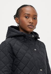 Črna prešita puhovka z kapuco, z zapenjanjem na pritisne gumbe in gladko, sijočo teksturo. Prešita z značilnim diamantnim vzorcem po celotni jakni.