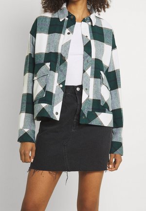 Personne portant une veste à carreaux verts et blancs boutonnée, un haut blanc et une jupe en jean noire avec ourlet brut.