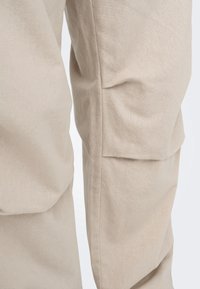 Beżowe spodnie bojówki z teksturowanego materiału, o luźnym kroju, z wieloma kieszeniami i widocznym przeszyciem wzdłuż szwów.