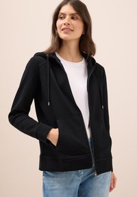 Felpa nera con zip in tessuto strutturato, dotata di cappuccio, tasche frontali e coulisse regolabili, abbinata a una maglietta bianca e jeans azzurri chiari.