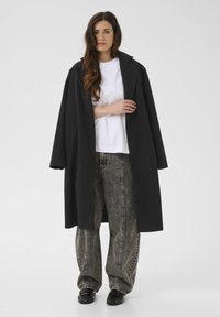 Manteau long noir avec grands revers, porté sur une chemise blanche. Associé à un jean gris ample taille haute et des chaussures noires.