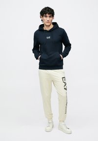 Felpa con cappuccio blu navy con tasca a canguro frontale e logo, abbinata a pantaloni jogger crema con stampa del logo laterale, indossata con sneakers bianche.
