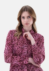 Blusa marrone con strisce a zebra rosa, caratterizzata da un collo alto e maniche lunghe con polsini a bottoni. Tessuto dalla texture liscia.